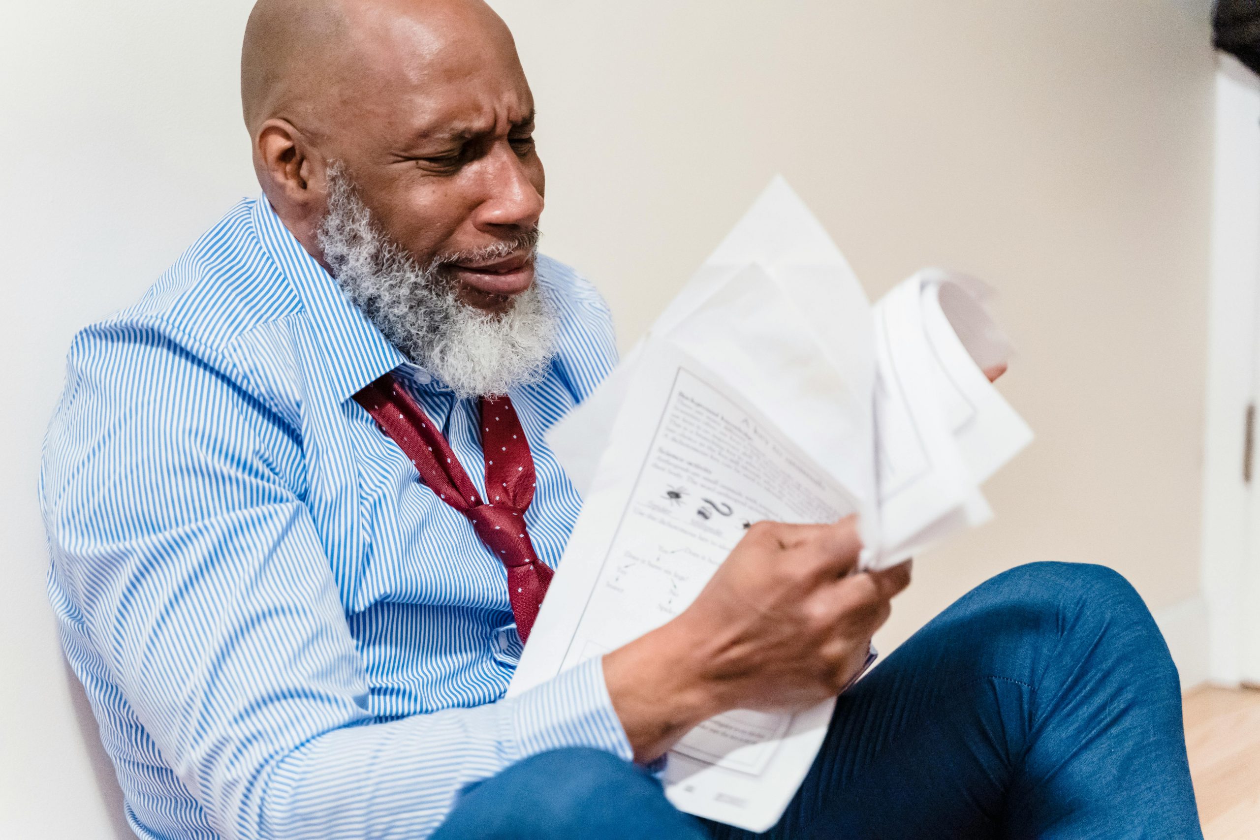 African American man in distress with paperwork, sitting against a wall indoors.